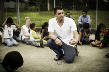 a man holding a rosary kneels down with children in a circle
